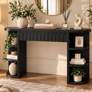 May include: A black console table with a curved top and arched detailing along the front apron. The piece features three open, rounded shelves on each side, providing storage for books, potted plants, a candle, and decorative objects.