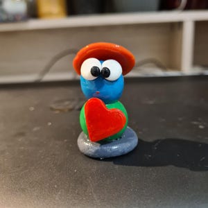 Peut inclure: Une figurine fantaisiste avec un corps bleu, de grands yeux blancs et un chapeau orange. Elle tient un cœur rouge contre une sphère verte, le tout reposant sur une base grise. La figurine a un design de dessin animé.