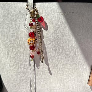 May include: A white pebbled leather tote bag with the text Tory Burch New York embossed on the front. A decorative bag charm hangs from the handle, featuring red, white, and translucent beads, a small red apple charm, and a braided tan cord.