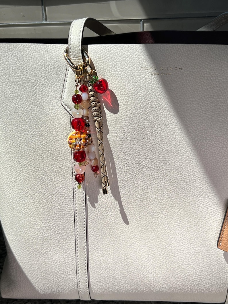 May include: A white pebbled leather tote bag with the text Tory Burch New York embossed on the front. A decorative bag charm hangs from the handle, featuring red, white, and translucent beads, a small red apple charm, and a braided tan cord.