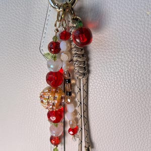 May include: A bag charm featuring a gold-tone lobster clasp, a braided beige cord with black flecks, and multiple dangling beads. The beads include red faceted spheres, translucent white spheres, and a textured orange-yellow round charm.