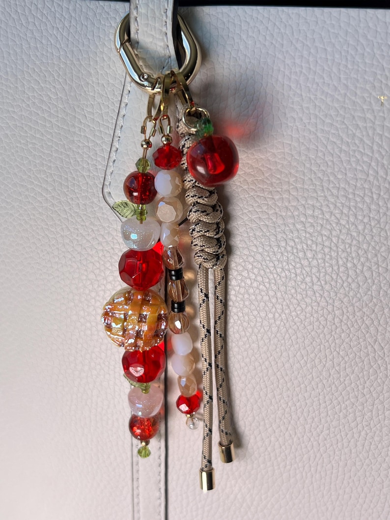 May include: A bag charm featuring a gold-tone lobster clasp, a braided beige cord with black flecks, and multiple dangling beads. The beads include red faceted spheres, translucent white spheres, and a textured orange-yellow round charm.