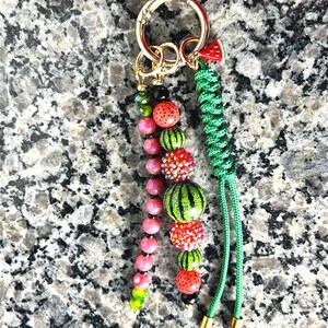 K&ouml;nnte beinhalten: Ein Schl&uuml;sselanh&auml;nger mit einem goldfarbenen Metallring und drei Anh&auml;ngern: eine Kette aus rosa und gr&uuml;nen Perlen, eine Kette aus wassermelonenfarbenen und glitzernden roten Perlen sowie eine gr&uuml;ne geflochtene Kordel mit einem kleinen Wassermelonen-Anh&auml;nger.