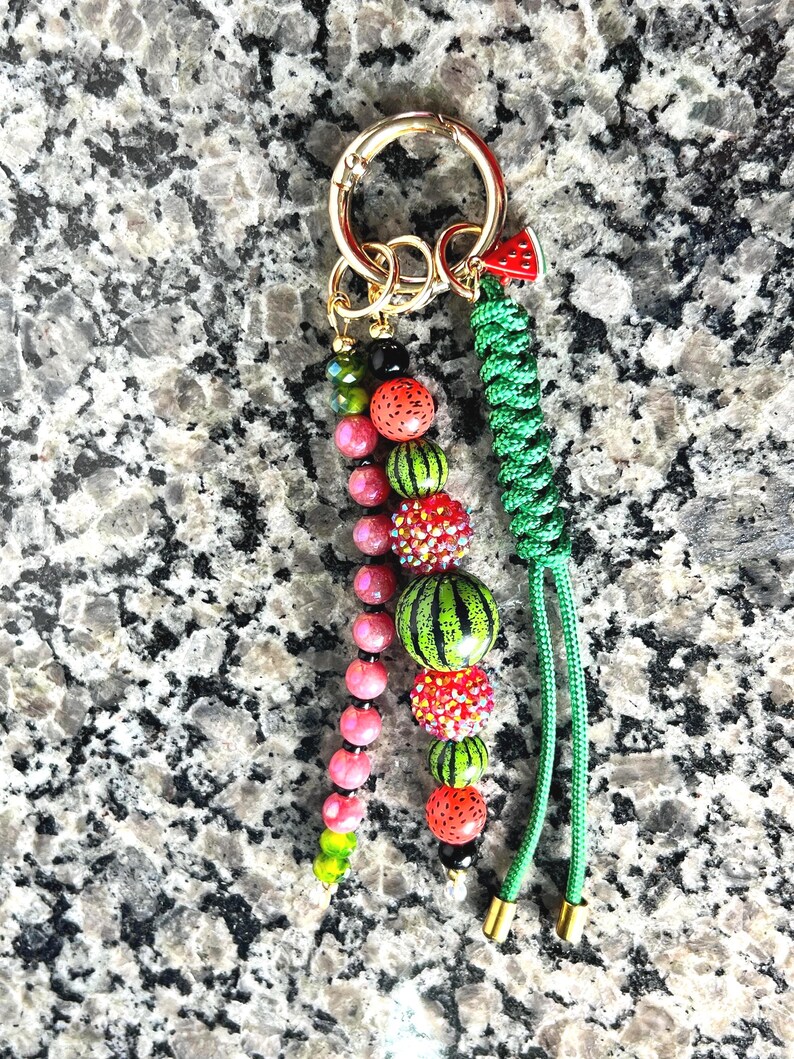 K&ouml;nnte beinhalten: Ein Schl&uuml;sselanh&auml;nger mit einem goldfarbenen Metallring und drei Anh&auml;ngern: eine Kette aus rosa und gr&uuml;nen Perlen, eine Kette aus wassermelonenfarbenen und glitzernden roten Perlen sowie eine gr&uuml;ne geflochtene Kordel mit einem kleinen Wassermelonen-Anh&auml;nger.