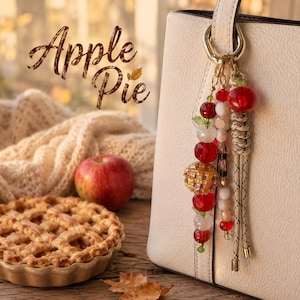 May include: A bag charm featuring red, white, and amber-colored beads, a small apple-shaped charm, and a braided tan cord, attached to a cream-colored handbag. The text "Apple Pie" is visible in the upper left corner above an apple and a pie.