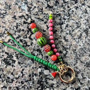 K&ouml;nnte beinhalten: Ein Schl&uuml;sselanh&auml;nger mit einer gr&uuml;nen geflochtenen Kordel, einem goldfarbenen Metallring und zwei Perlenstr&auml;ngen. Ein Strang hat wassermelonenf&ouml;rmige und glitzernde rote Perlen, der andere rosa und gr&uuml;ne Perlen. Ein kleiner Wassermelonen-Anh&auml;nger.