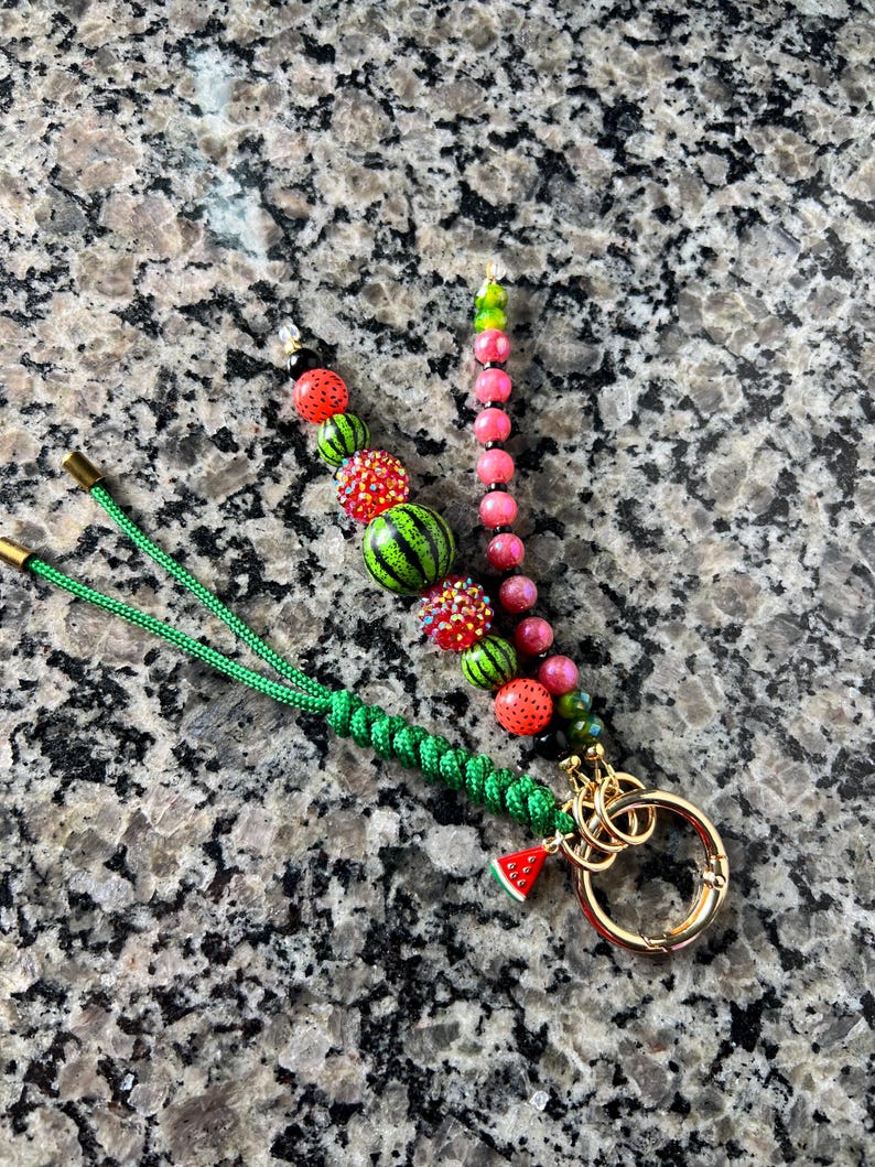 K&ouml;nnte beinhalten: Ein Schl&uuml;sselanh&auml;nger mit einer gr&uuml;nen geflochtenen Kordel, einem goldfarbenen Metallring und zwei Perlenstr&auml;ngen. Ein Strang hat wassermelonenf&ouml;rmige und glitzernde rote Perlen, der andere rosa und gr&uuml;ne Perlen. Ein kleiner Wassermelonen-Anh&auml;nger.