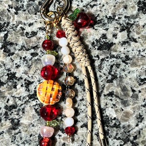 May include: A keychain featuring a gold-toned lobster clasp with two beaded strands and a braided tan cord. One strand includes red, white, and iridescent beads with a miniature orange pie charm. The other strand has clear, white, and red beads.