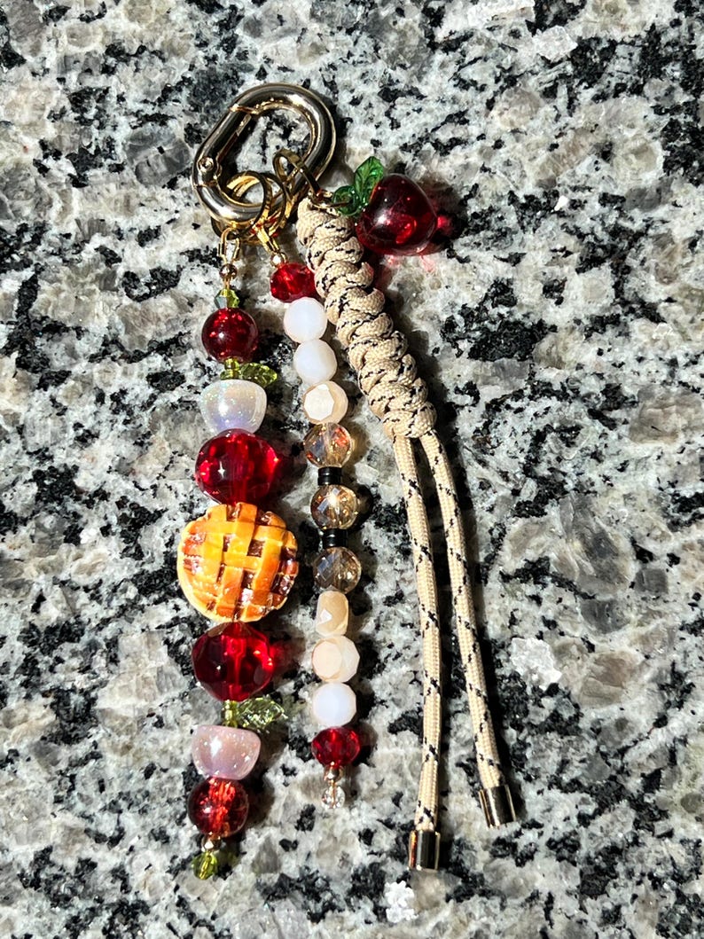 May include: A keychain featuring a gold-toned lobster clasp with two beaded strands and a braided tan cord. One strand includes red, white, and iridescent beads with a miniature orange pie charm. The other strand has clear, white, and red beads.