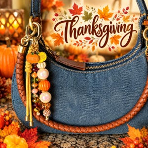 May include: A denim handbag with a braided brown leather handle features a decorative charm dangling from a gold-toned ring. The charm includes yellow cord tassels, orange pumpkin-shaped beads, and various multicolored glass and pearl beads.