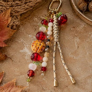 May include: A keychain featuring a gold-toned lobster clasp, a braided beige cord with black accents, and two dangling strands of beads. The beads include red faceted spheres, white spheres, small green leaf charms, and a miniature round pie charm.