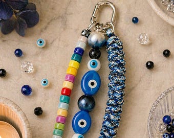 Może przedstawiać: Brelok do kluczy z motywem Evil Eye. Brelok posiada srebrny karabińczyk, kolorowe koraliki i niebieskie zawieszki Evil Eye. Tekst "Evil Eye" jest wyświetlany u góry obrazu.