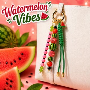A bag charm featuring a gold-toned ring with dangling strands of pink, green, and black beads, textured watermelon-patterned spheres, and a braided green cord. A small watermelon charm is attached. The text Watermelon Vibes is visible.