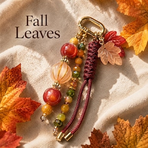May include: A keychain featuring the text Fall Leaves, adorned with a gold-toned metal clasp, a dark red braided cord, and a collection of beads in shades of red, orange, and green, alongside two translucent leaf-shaped charms.