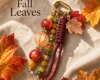 Może przedstawiać: Brelok z napisem Fall Leaves, ozdobiony złotym metalowym zapięciem, ciemnoczerwonym plecionym sznurkiem oraz kolekcją koralików w odcieniach czerwieni, pomarańczu i zieleni, wraz z dwoma półprzezroczystymi zawieszkami w kształcie liści.