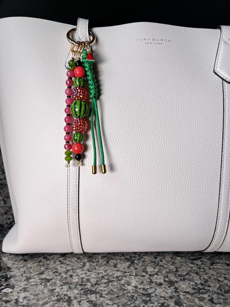 K&ouml;nnte beinhalten: Eine wei&szlig;e Ledertasche mit einem goldfarbenen Logo mit der Aufschrift Tory Burch New York. Am Griff befindet sich ein dekorativer Anh&auml;nger mit einer Reihe mehrfarbiger Perlen in Rosa, Gr&uuml;n und Schwarz sowie einer gr&uuml;nen geflochtenen Kordel.