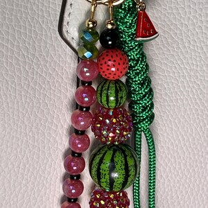 K&ouml;nnte beinhalten: Ein dekorativer Taschenanh&auml;nger mit drei Str&auml;ngen an goldfarbenen Ringen. Ein Strang besteht aus facettierten rosa und gr&uuml;nen Perlen, ein weiterer aus glitzernden rot-gr&uuml;nen Perlen und Wassermelonen-Motiven, und der dritte ist eine geflochtene gr&uuml;ne Kordel mit einem Wassermelonen-Anh&auml;nger.