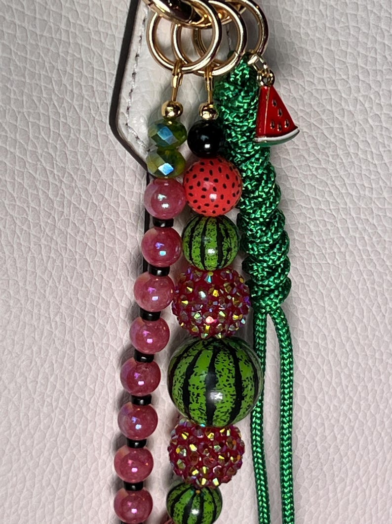 K&ouml;nnte beinhalten: Ein dekorativer Taschenanh&auml;nger mit drei Str&auml;ngen an goldfarbenen Ringen. Ein Strang besteht aus facettierten rosa und gr&uuml;nen Perlen, ein weiterer aus glitzernden rot-gr&uuml;nen Perlen und Wassermelonen-Motiven, und der dritte ist eine geflochtene gr&uuml;ne Kordel mit einem Wassermelonen-Anh&auml;nger.