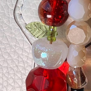 May include: A beaded accessory featuring a faceted red sphere, a translucent white apple-shaped bead with iridescent glitter, and a small green leaf-shaped bead. Beside it is a strand of faceted white and clear beads with a dark base component.
