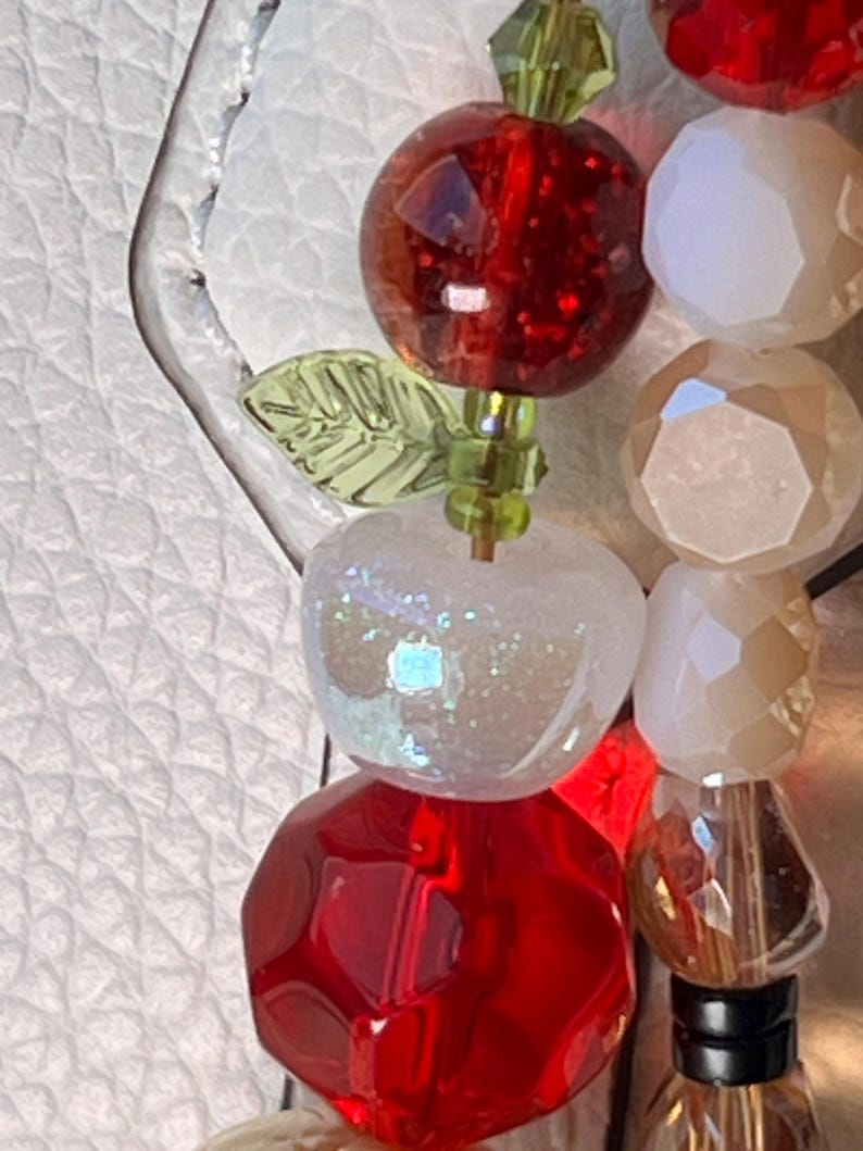 May include: A beaded accessory featuring a faceted red sphere, a translucent white apple-shaped bead with iridescent glitter, and a small green leaf-shaped bead. Beside it is a strand of faceted white and clear beads with a dark base component.