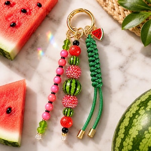 May include: A gold-toned metal keychain featuring three distinct dangling elements: a strand of pink and green beads, a central column of watermelon-patterned and sparkling red beads, and a braided green cord with a small watermelon charm.