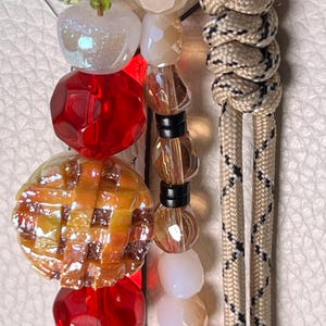 May include: A decorative beaded accessory featuring a central charm shaped like a lattice-topped pie, surrounded by faceted red beads, translucent white beads, and a beige paracord lanyard with a black and white diamond pattern woven throughout.