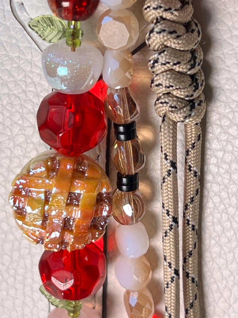 May include: A decorative beaded accessory featuring a central charm shaped like a lattice-topped pie, surrounded by faceted red beads, translucent white beads, and a beige paracord lanyard with a black and white diamond pattern woven throughout.