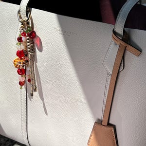 May include: A white pebbled leather tote bag with the Tory Burch logo embossed on the front. A decorative charm featuring red, orange, and clear beads, a small cherry-shaped pendant, and braided tan cords hangs from one of the top handles.