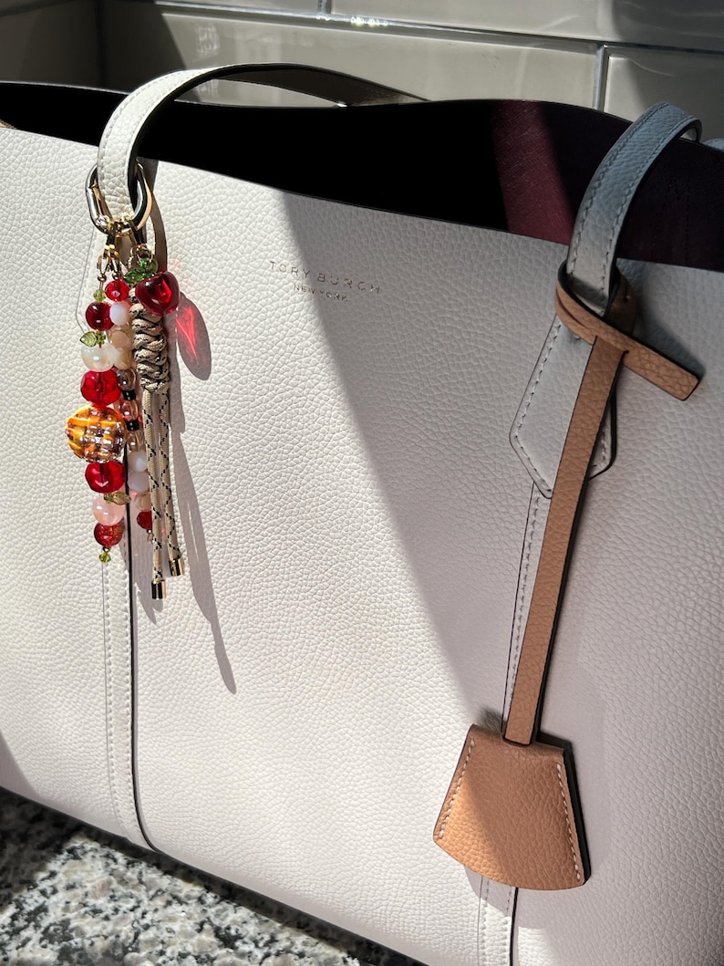 May include: A white pebbled leather tote bag with the Tory Burch logo embossed on the front. A decorative charm featuring red, orange, and clear beads, a small cherry-shaped pendant, and braided tan cords hangs from one of the top handles.