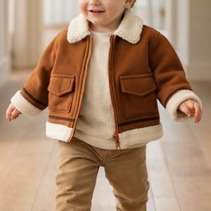 Peut inclure: Un jeune enfant vêtu d'une veste marron avec un col et des poignets en fausse fourrure crème, associé à un pantalon et des chaussures beiges. La veste a deux poches avant et une fermeture éclair. L'enfant marche sur un sol en bois.