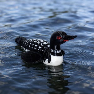 May include: A 3D printed loon figurine featuring a black and white checkered pattern on its back, a solid black head and neck, a white chest, and a sharp black beak with a distinct red eye detail, floating on a rippling blue water surface.