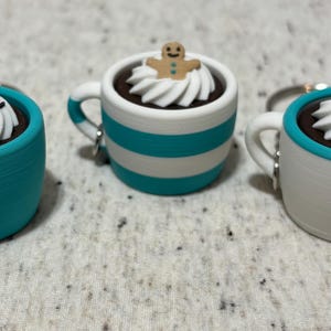 Puede incluir: Tres llaveros en miniatura con forma de tazas de café. Uno es turquesa, otro blanco con rayas turquesas y otro blanco con borde turquesa. Cada taza está llena de una sustancia marrón oscuro y cubierta con crema batida blanca.