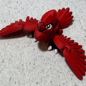 May include: A bright red cardinal figurine with outstretched wings. The bird has a detailed design, including visible feathers and a cartoonish face with white eyes and a black outline. The figurine is a vibrant red color.