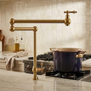 Peut inclure: Un robinet remplisseur de casserole mural avec une finition en laiton brossé, doté d'un bras articulé à double articulation et d'une poignée à levier unique. Le robinet est placé au-dessus d'une marmite bleu foncé avec des poignées cuivrées.