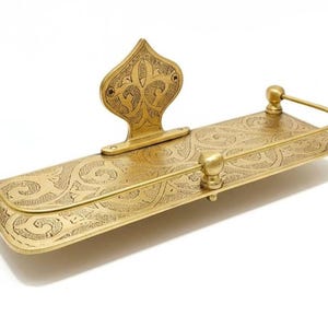May include: A brass-colored shelf with an ornate, engraved design. The shelf has a rectangular base with rounded corners and a decorative back piece. The design includes floral and scroll patterns. The shelf is likely for decorative or functional use.