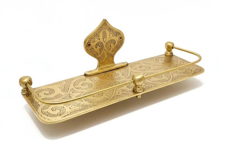 May include: A brass-colored shelf with an ornate, engraved design. The shelf has a rectangular base with rounded corners and a decorative back piece. The design includes floral and scroll patterns. The shelf is likely for decorative or functional use.