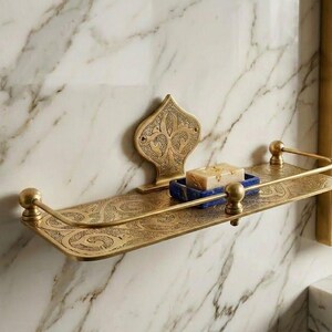 May include: Ornate brass bathroom shelf with a decorative backplate and curved front rail. The shelf holds a blue soap dish with a bar of soap. The shelf is mounted on a marble wall.