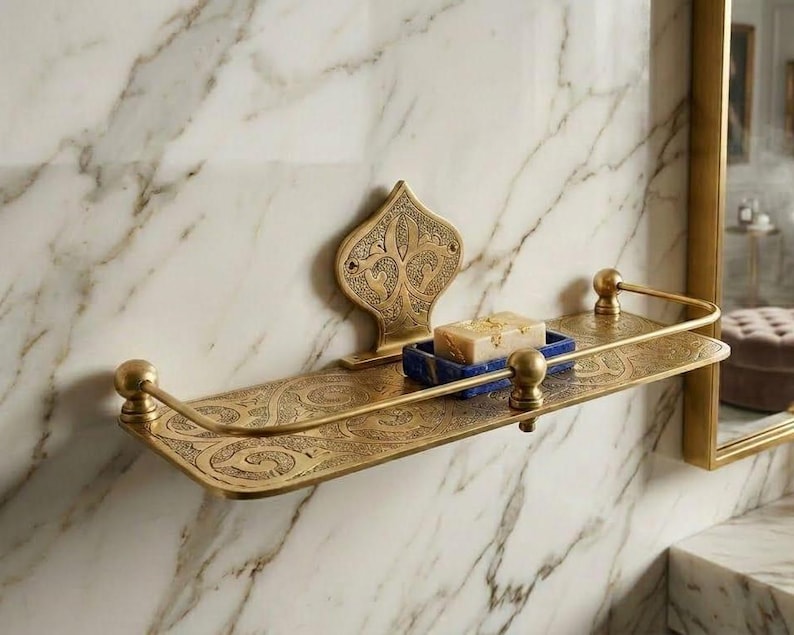 May include: Ornate brass bathroom shelf with a decorative backplate and curved front rail. The shelf holds a blue soap dish with a bar of soap. The shelf is mounted on a marble wall.