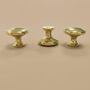 May include: Three polished gold-tone metal cabinet knobs with a smooth, rounded top surface and a flared base. Each knob features a reflective, high-gloss finish and a central circular opening on the top, showcasing a uniform, metallic design.