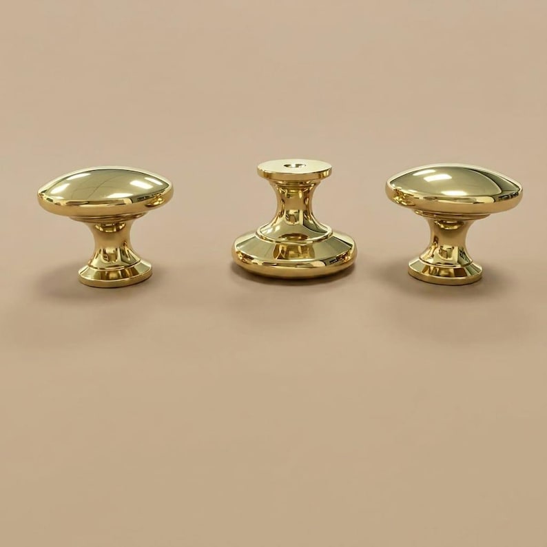 May include: Three polished gold-tone metal cabinet knobs with a smooth, rounded top surface and a flared base. Each knob features a reflective, high-gloss finish and a central circular opening on the top, showcasing a uniform, metallic design.