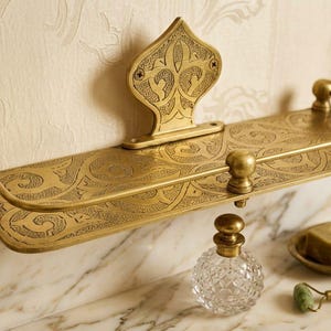 May include: Ornate brass bathroom shelf with an etched floral design. The shelf has a decorative backplate and rounded end supports. A clear glass perfume bottle, a soap dish, and a jade roller are on the marble surface.
