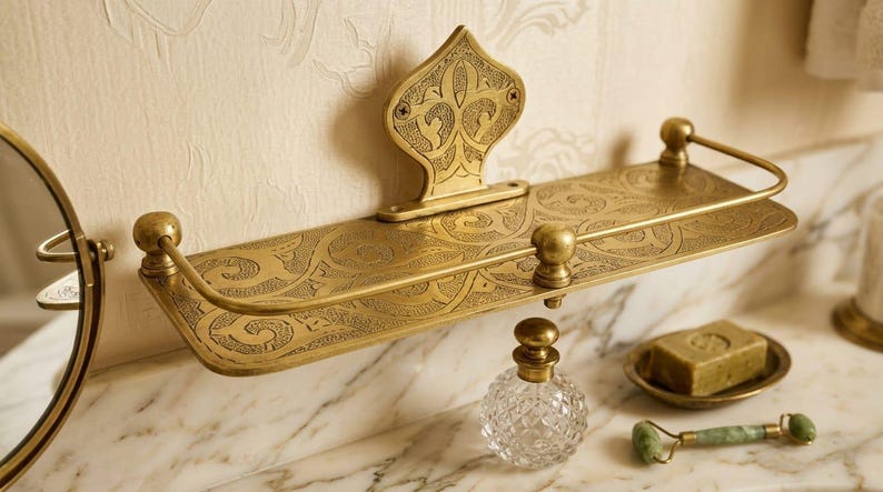May include: Ornate brass bathroom shelf with an etched floral design. The shelf has a decorative backplate and rounded end supports. A clear glass perfume bottle, a soap dish, and a jade roller are on the marble surface.