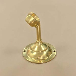 May include: A polished brass curtain rod holder. It features a circular base with two screw holes and a curved arm to hold the rod. The brass has a shiny, reflective finish. The holder is designed for mounting.