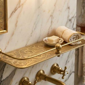 May include: A decorative brass shelf with an ornate, engraved design. The shelf holds a rolled-up beige towel, a small soap dish with a bar of soap, and is mounted on a marble wall. The shelf has a curved front rail and ball finials.