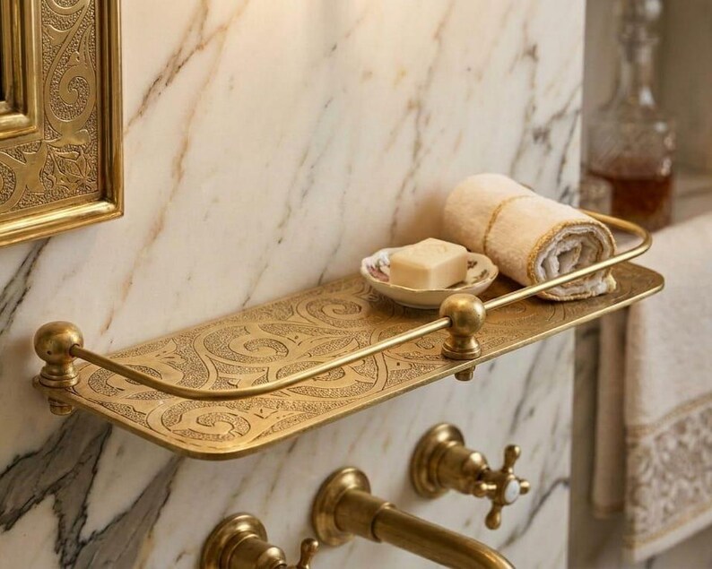 May include: A decorative brass shelf with an ornate, engraved design. The shelf holds a rolled-up beige towel, a small soap dish with a bar of soap, and is mounted on a marble wall. The shelf has a curved front rail and ball finials.