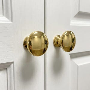 May include: Two polished brass cabinet knobs with a smooth, rounded, dome-shaped design. Each knob features a reflective, metallic gold-toned finish and is mounted onto a white wooden cabinet door with a visible grain texture and panel detailing.