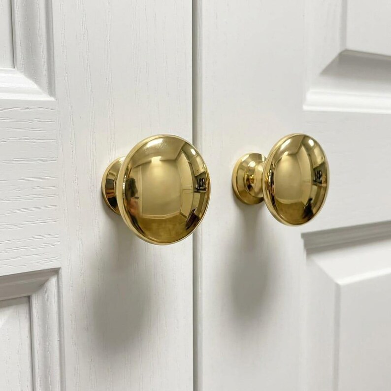 May include: Two polished brass cabinet knobs with a smooth, rounded, dome-shaped design. Each knob features a reflective, metallic gold-toned finish and is mounted onto a white wooden cabinet door with a visible grain texture and panel detailing.