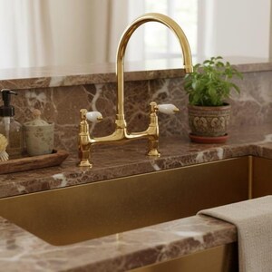 Könnte beinhalten: Ein goldfarbener Küchenhahn mit weißen Griffen ist auf einer braunen Marmorarbeitsplatte montiert. Eine kleine Topfpflanze befindet sich rechts vom Wasserhahn. Ein goldfarbenes Waschbecken ist darunter zu sehen.