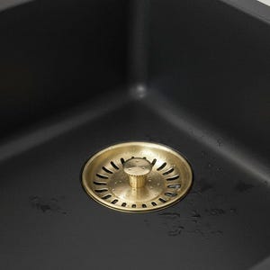 May include: A close-up shot of a black kitchen sink featuring a gold-coloured drain. The sink is square and has a matte finish. The drain is circular with small holes and a central knob.