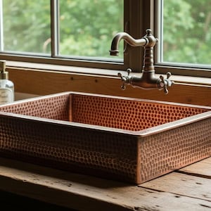 May include: A copper rectangular sink with a hammered texture sits on a wooden surface. A vintage-style faucet and a small succulent in a white pot are nearby. A stack of folded towels and a soap dispenser are also visible.